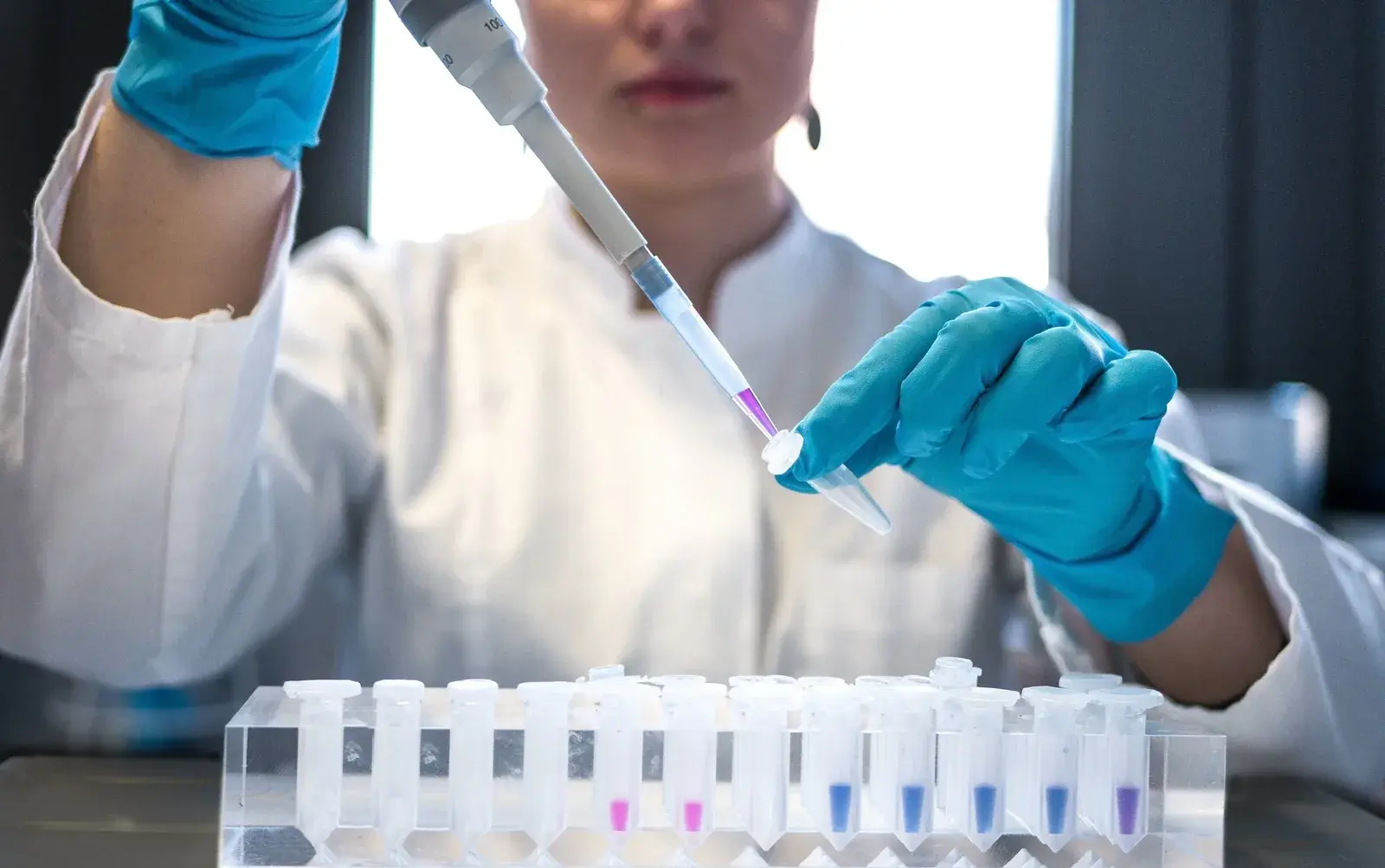A researcher in a white lab coat and blue nitrile gloves dispensing liquid from a pipette into a microcentrifuge tube, over a rack of tubes holding pink, blue and purple samples.